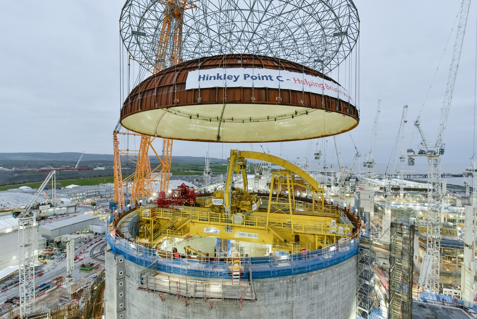Hinkley Point C reactor building dome being lifted into place by crane.